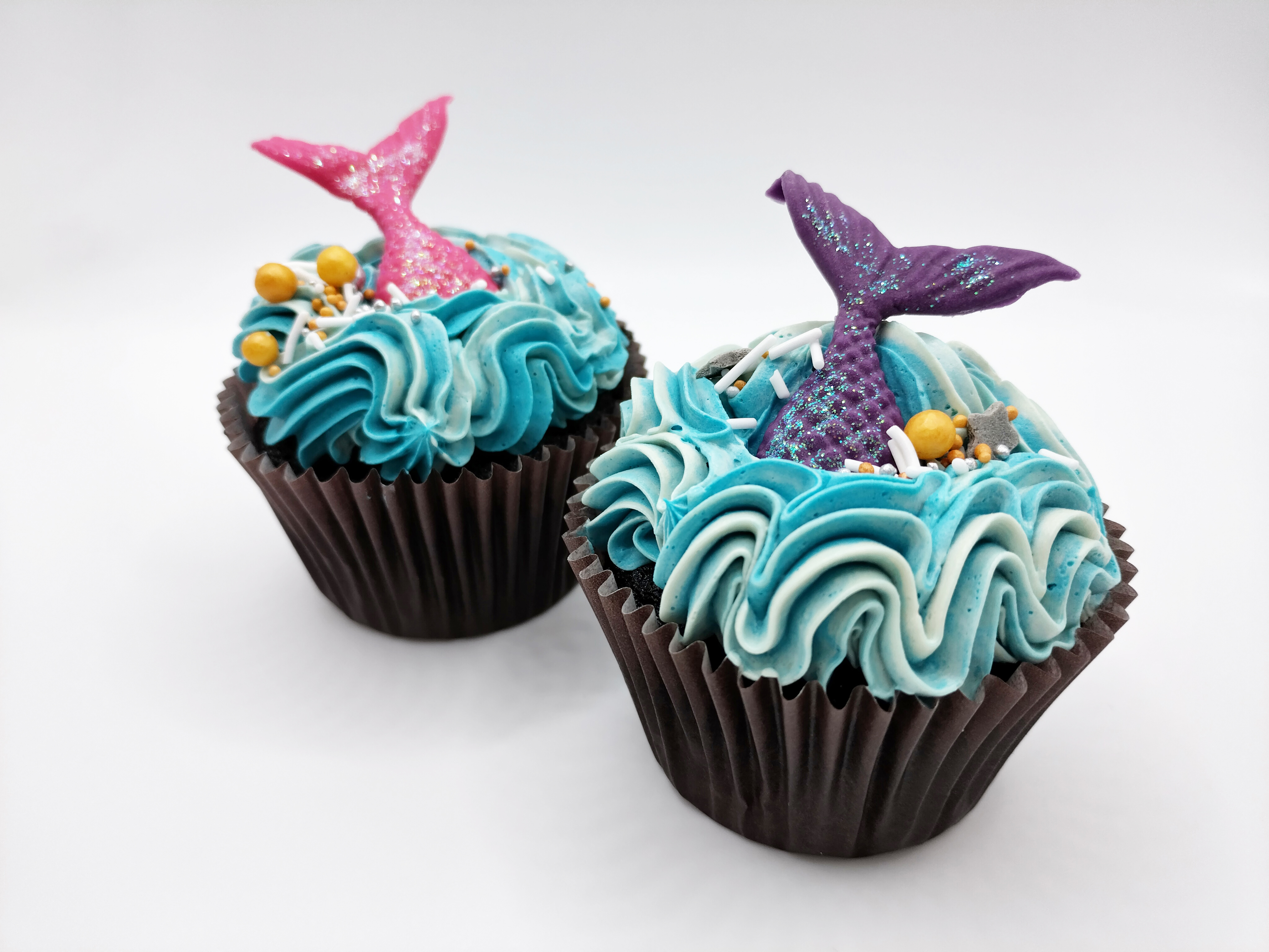 Two mermaid themed cupcakes, blue wavy buttercream, shiny sugar pearls and pink and purple mermaid tails
